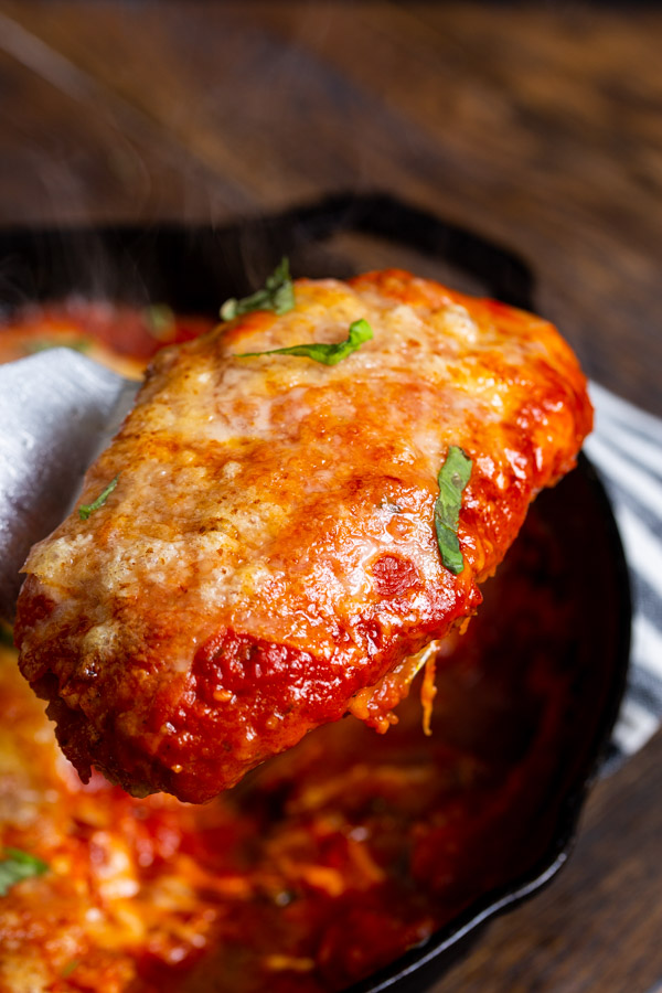 A close-up of a piece of cheesy chicken parmesan being lifted from a skillet with a spatula.