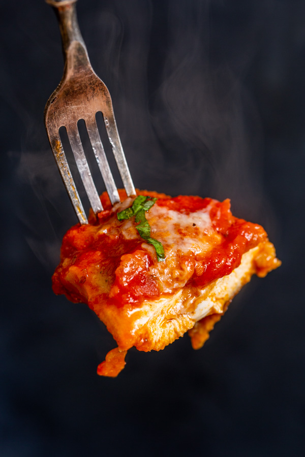 A silver fork holds a steaming piece of cheesy, saucy chicken with a garnish of fresh basil.