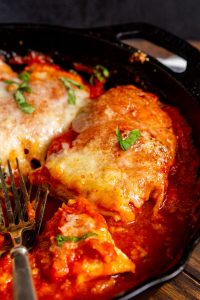 A close-up of chicken parmesan in a skillet, featuring breaded chicken cutlets topped with marinara sauce, melted cheese, and garnished with basil leaves. A fork is placed at the bottom left.