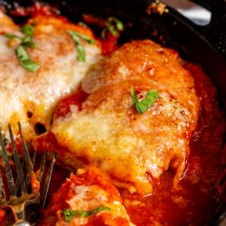 A close-up of chicken parmesan in a skillet, featuring breaded chicken cutlets topped with marinara sauce, melted cheese, and garnished with basil leaves. A fork is placed at the bottom left.