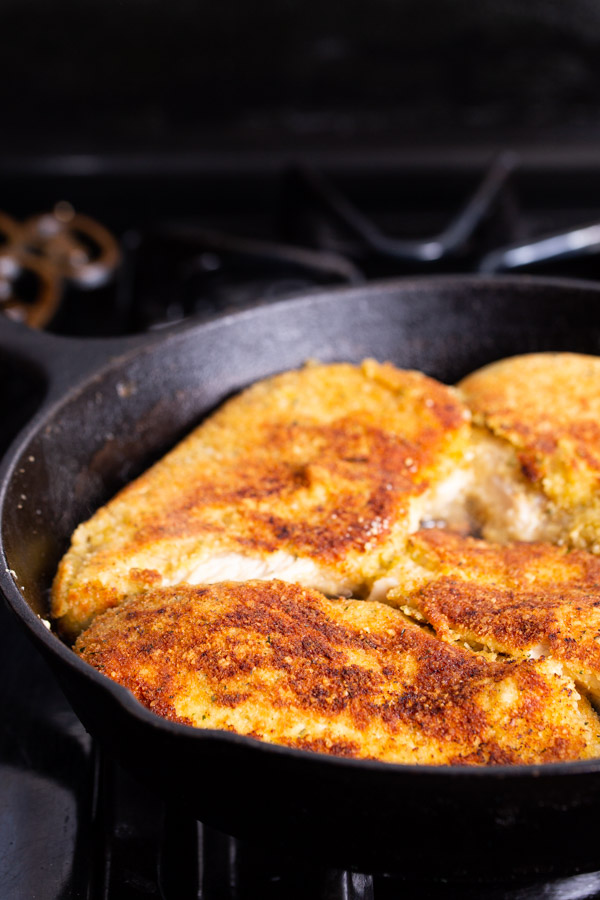 A close-up of golden-brown breaded chicken breasts cooking in a black cast iron skillet on a stovetop.