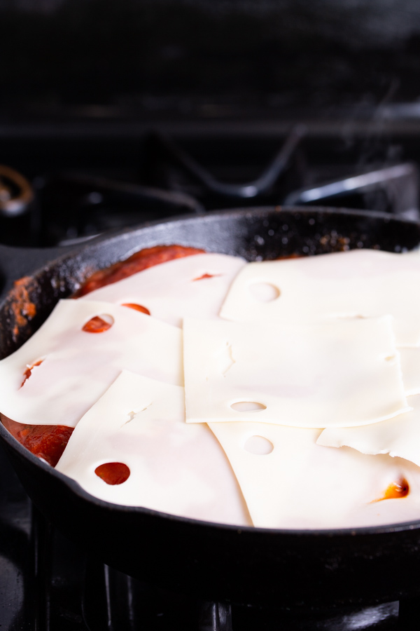 Slices of Swiss cheese melting over meat in a black skillet on a stovetop.