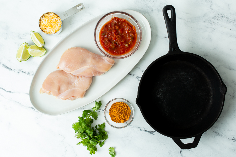 Top-down view of ingredients for a chicken dish including raw chicken breasts, cheese, salsa, lime wedges, seasoning, cilantro, and an empty cast-iron skillet on a marble countertop.