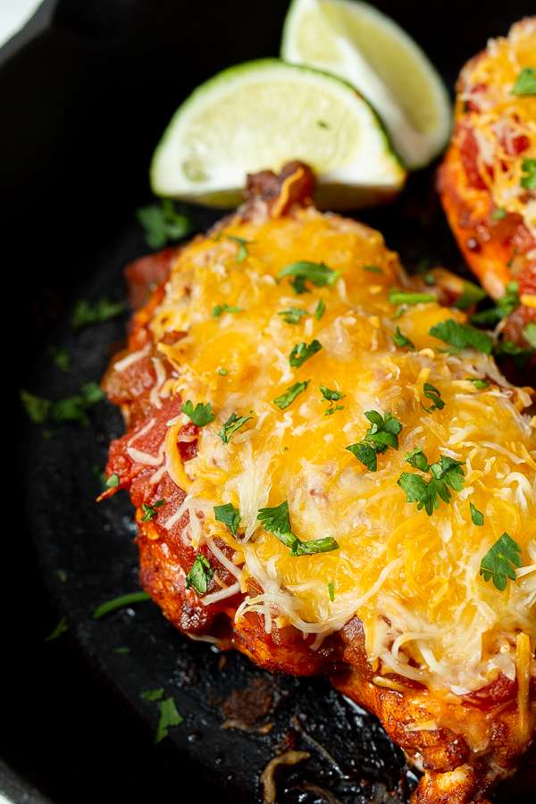 Close-up of a cooked dish featuring a cheese-topped chicken breast garnished with chopped parsley and served with lime wedges.