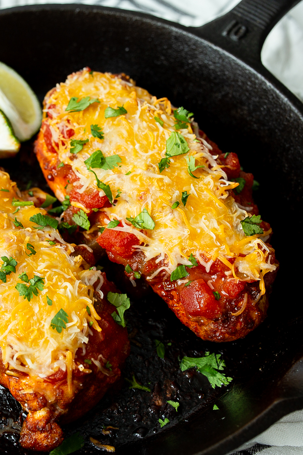 Two pieces of chicken in a skillet, topped with melted cheese, tomato sauce, and chopped cilantro, with lime wedges in the background.