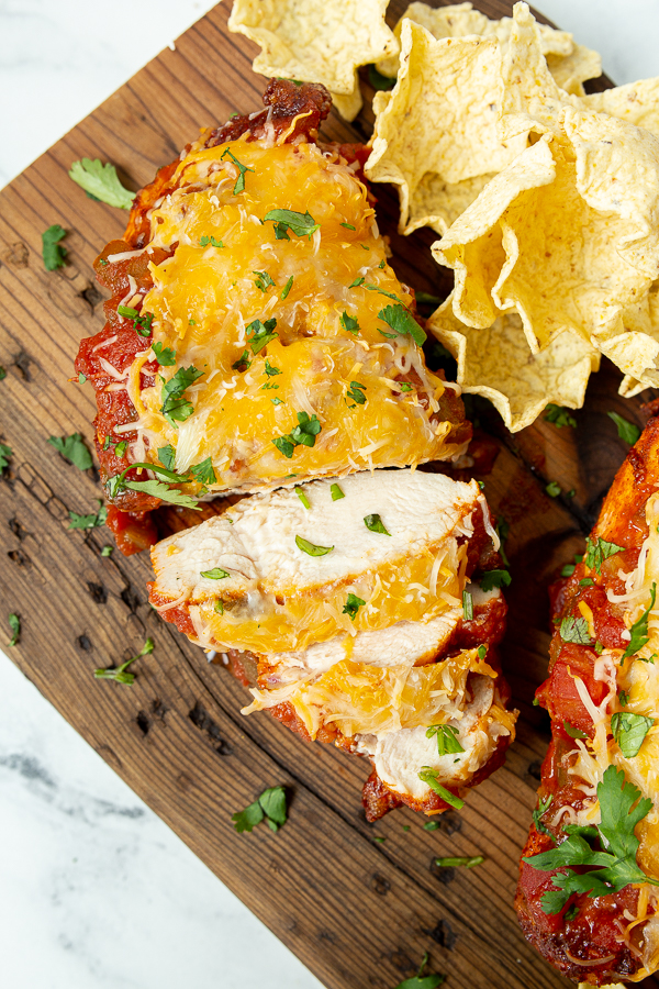 A wooden board holding a sliced cheese-covered chicken breast garnished with parsley, next to a serving of tortilla chips.