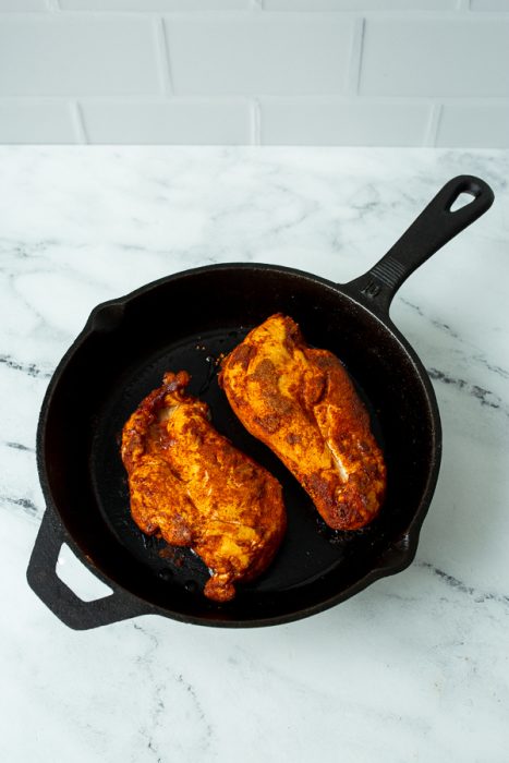 Two seasoned chicken breasts in a black cast iron skillet on a marble countertop with a white tile background.