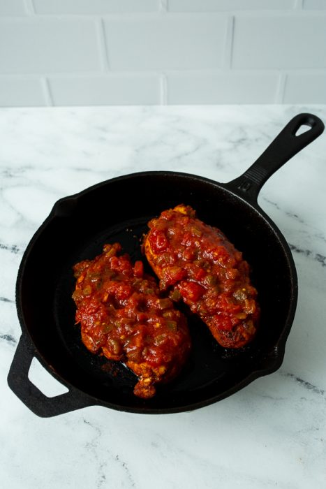 Two pieces of chicken topped with tomato sauce and diced vegetables cooking in a black cast iron skillet on a white marble countertop.