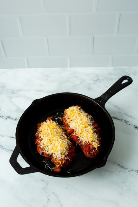 Two cooked chicken breasts topped with melted cheese in a black cast iron skillet, placed on a marble counter.