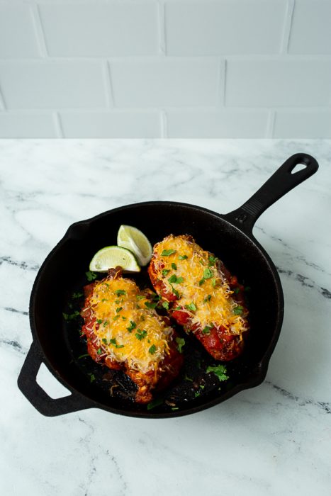 Two cheese-topped chicken breasts baked in a black skillet, garnished with chopped herbs and served with lime wedges on a marble countertop.