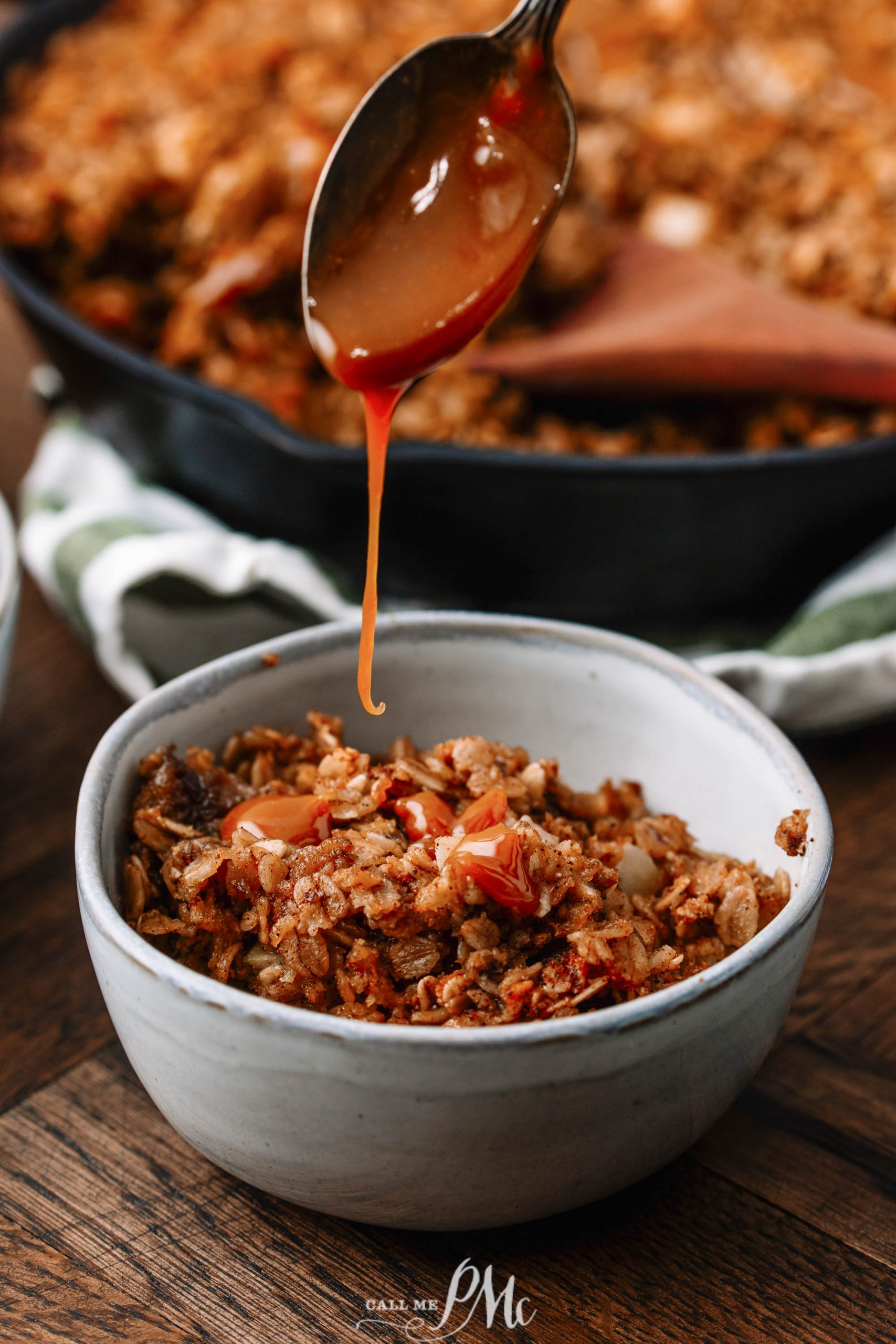 A bowl of baked oatmeal with fruit on a wooden table, as caramel sauce is being spooned on top. A pan with more oatmeal and a wooden spoon are visible in the background.