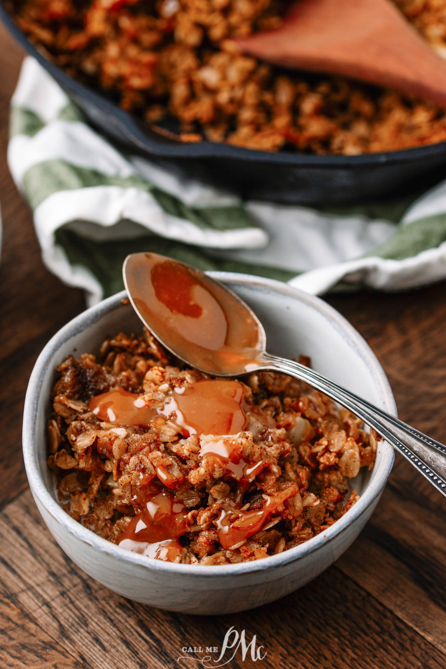 A bowl of apple crisp topped with caramel sauce. A spoon rests on the edge of the bowl, and a pan of the dessert is visible in the background.