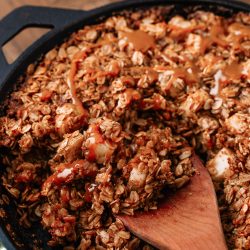 A close-up of a baked dish in a cast iron skillet, with a wooden spoon stirring a mixture that appears to be an oatmeal-based recipe drizzled with a sauce.