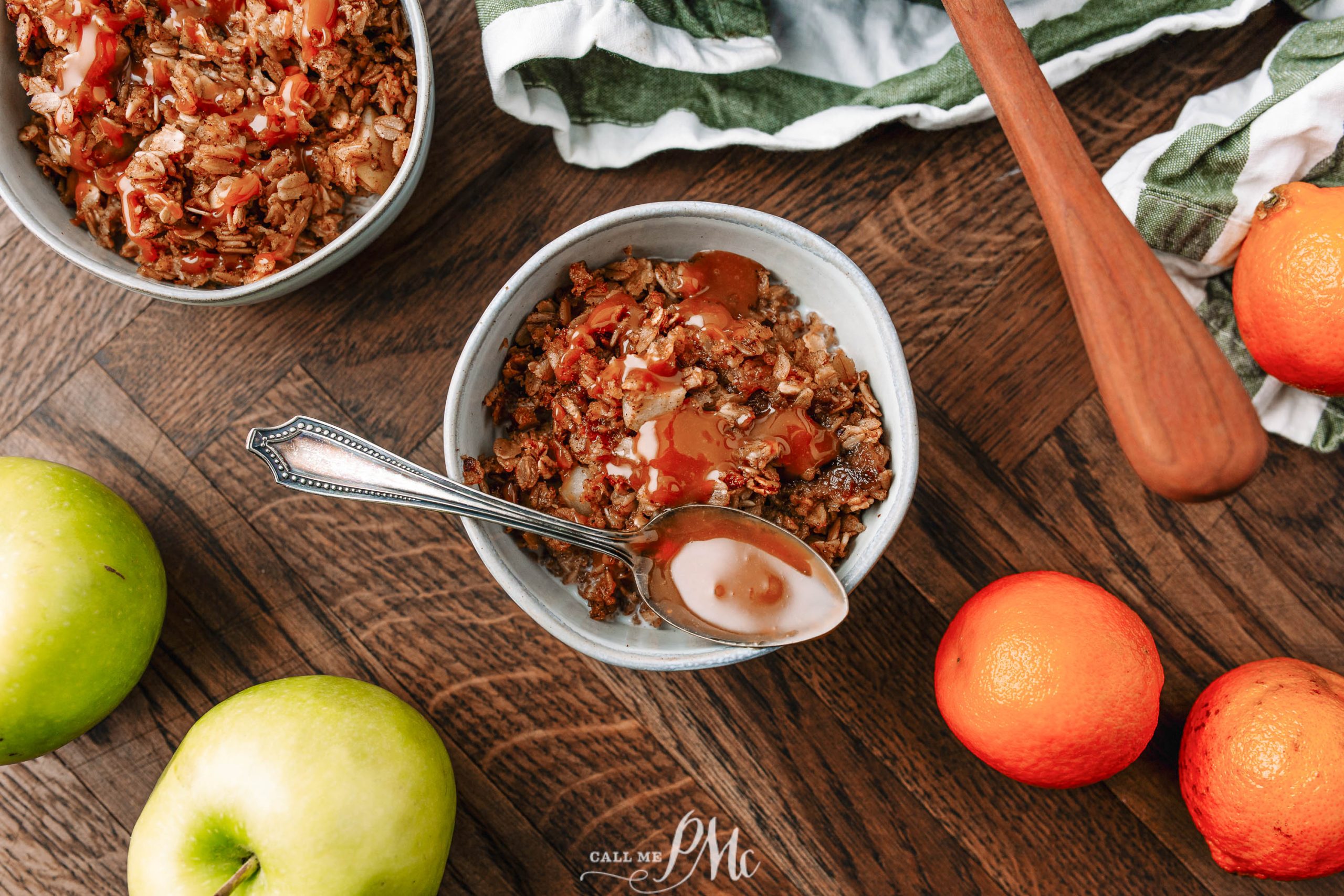 A bowl of granola with a spoon is placed on a wooden surface, surrounded by apples, oranges, another bowl of granola, and a wooden utensil. A patterned cloth is partially visible in the background.