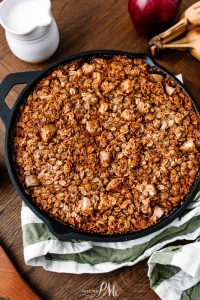 A cast iron skillet filled with baked oatmeal rests on a green-striped kitchen towel. A jug of milk, a red apple, and bananas are in the background on a wooden surface.