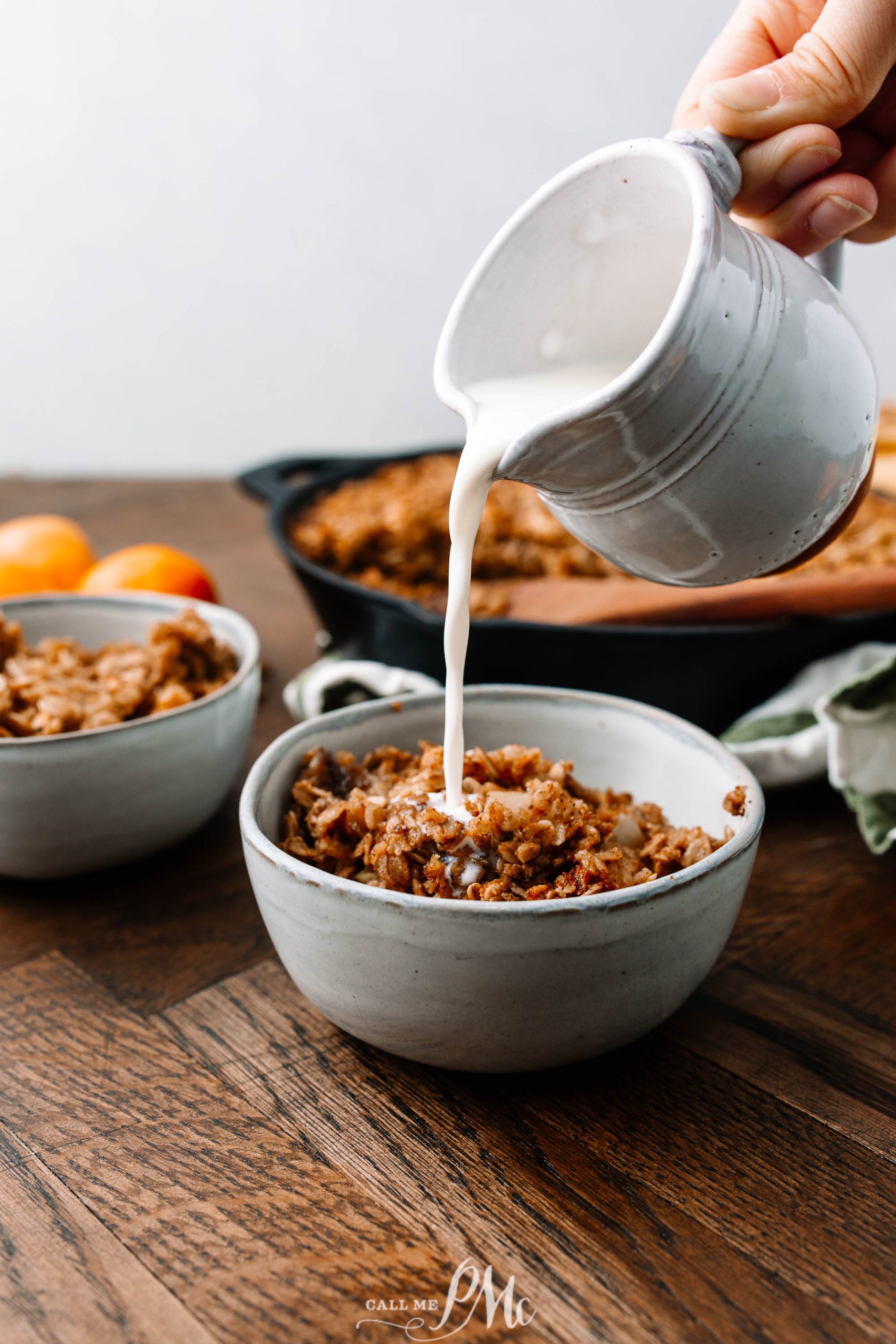 A person is pouring milk from a jug into a bowl of cereal on a wooden table. Other bowls and food are visible in the background.