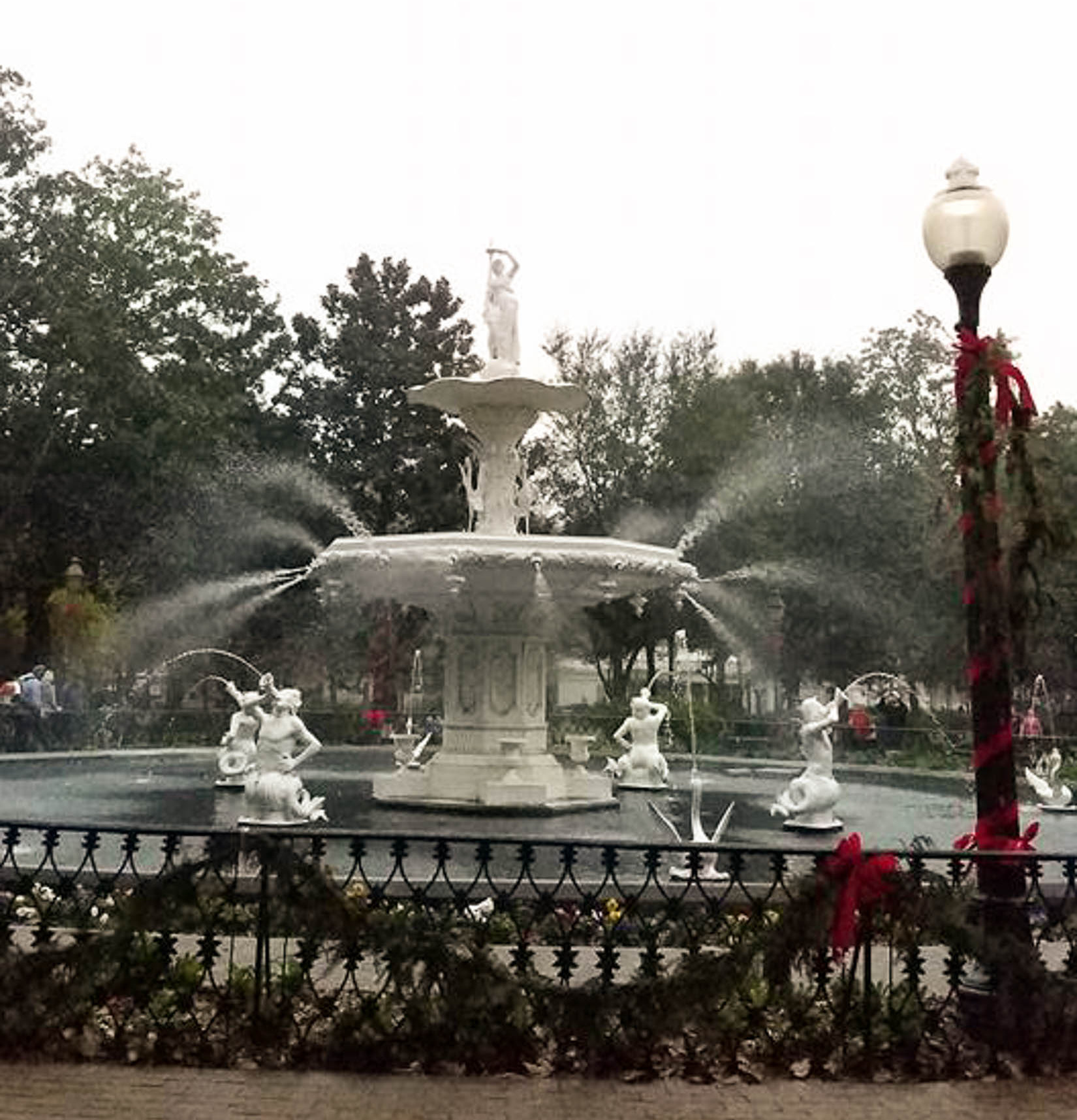 A white fountain with multiple water streams and statues surrounded by trees and a decorated fence with red ribbons.