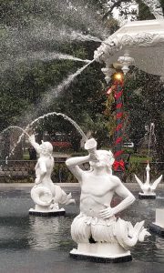 A water fountain with white, mermaid-like statues spouting water from their mouths into the air, surrounded by greenery.