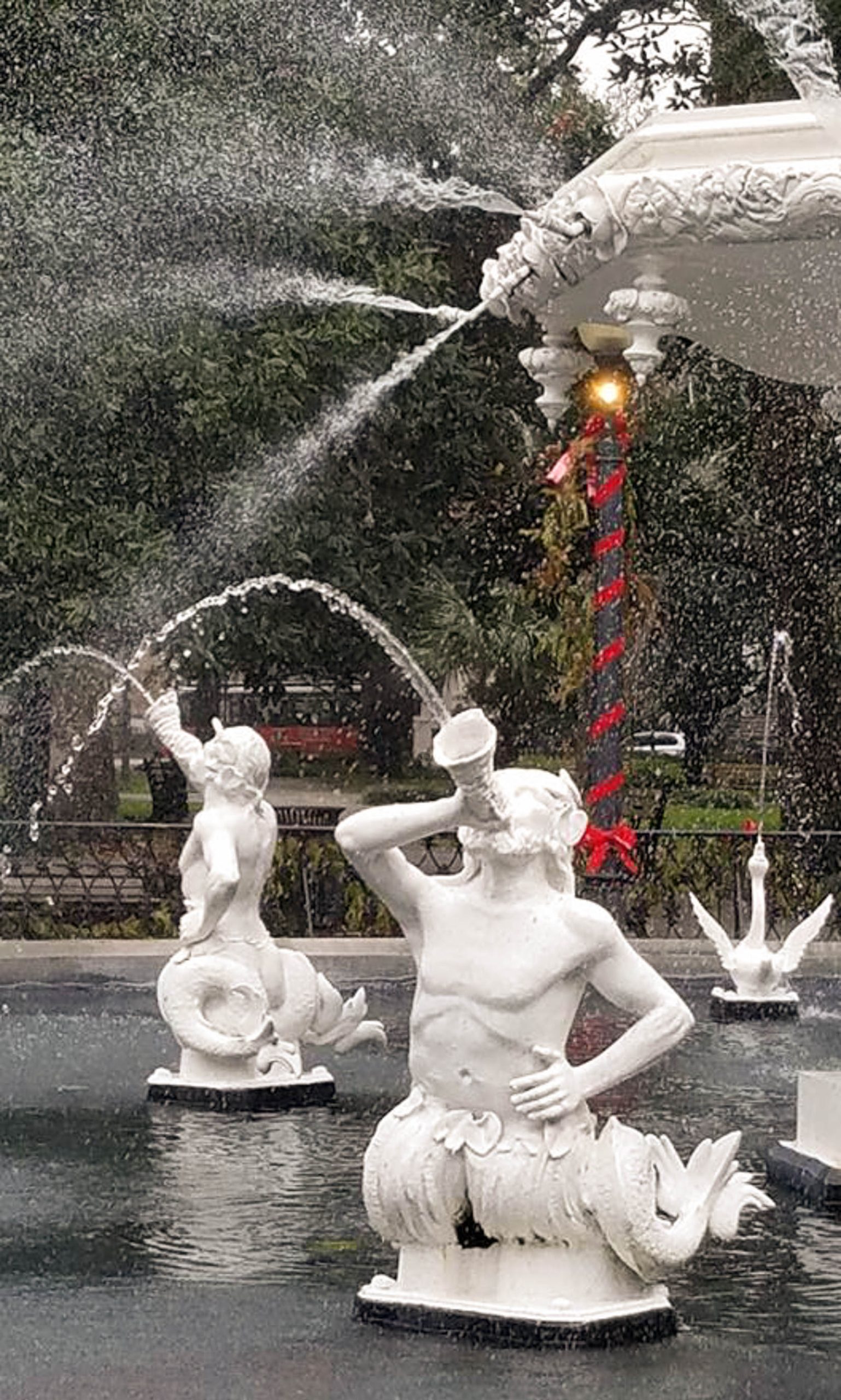 A water fountain with white, mermaid-like statues spouting water from their mouths into the air, surrounded by greenery.