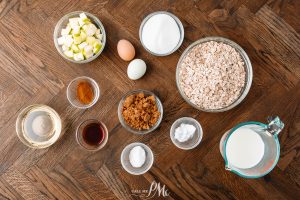 An arrangement of baking ingredients on a wooden surface includes chopped apples, oats, eggs, sugar, brown sugar, cinnamon, baking powder, salt, vanilla extract, oil, and milk.