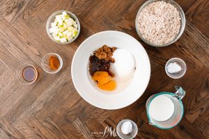 Overhead view of various baking ingredients in bowls on a wooden surface, including eggs, brown sugar, vanilla extract, and chopped apples.