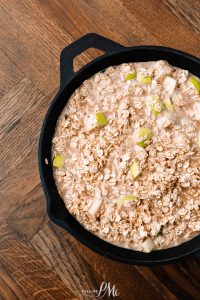 A black cast iron skillet filled with uncooked oatmeal mixed with diced apples, placed on a wooden table.