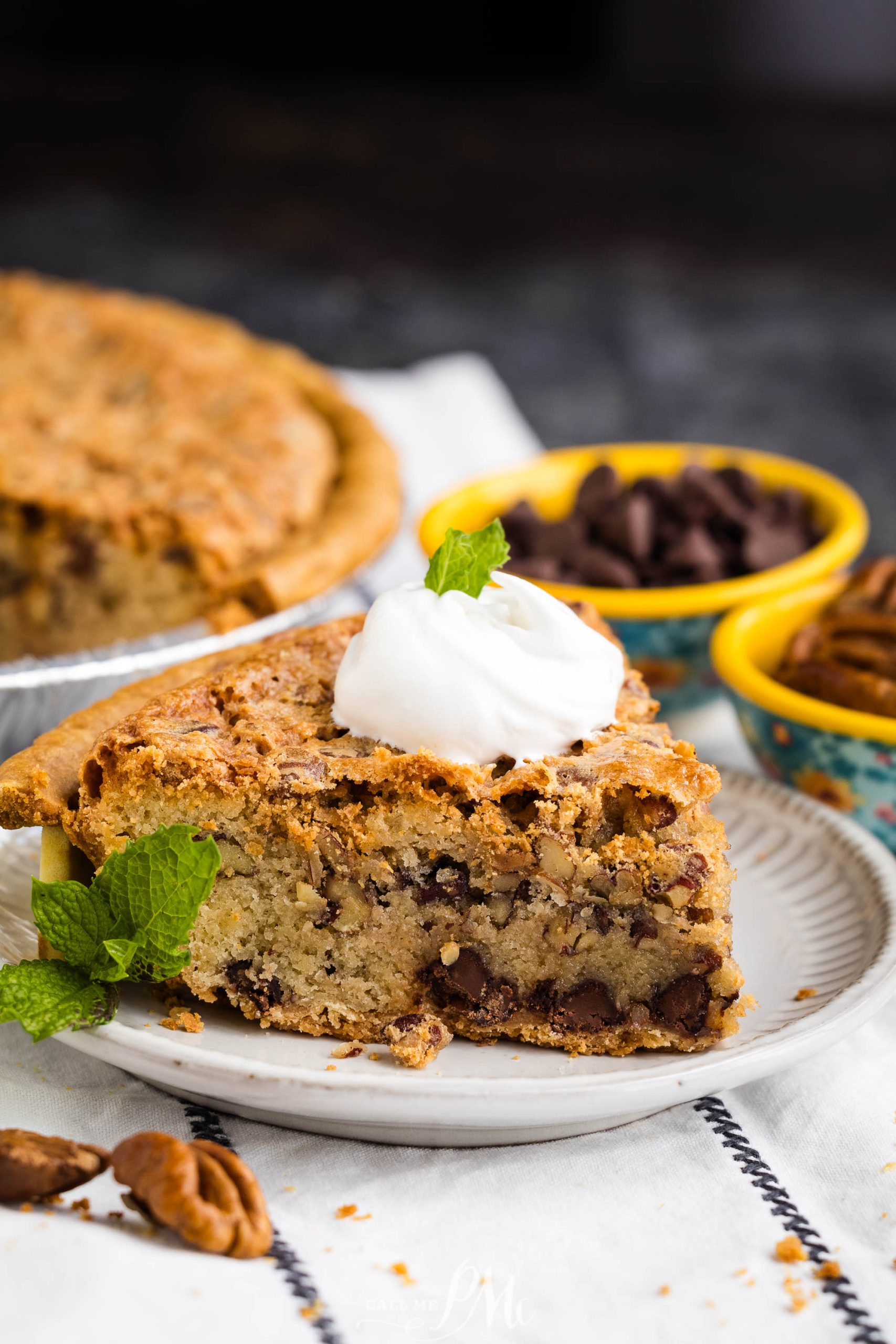 A slice of chocolate chip pecan pie topped with a dollop of whipped cream and garnished with a mint leaf, served on a white plate. Two bowls of chocolate chips and pecans are in the background.