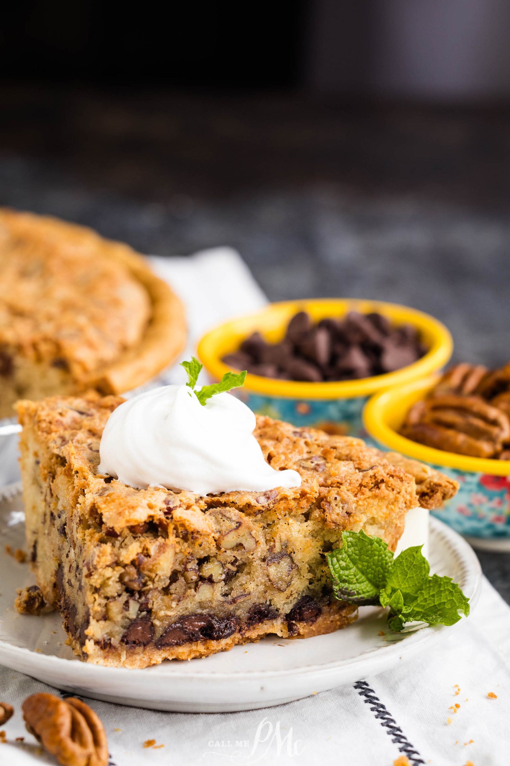 A slice of Kentucky Derby Pie with a crumbly crust, filled with chocolate chips and topped with whipped cream, is plated alongside bowls containing chocolate chips and pecans. A sprig of mint garnishes the plate.