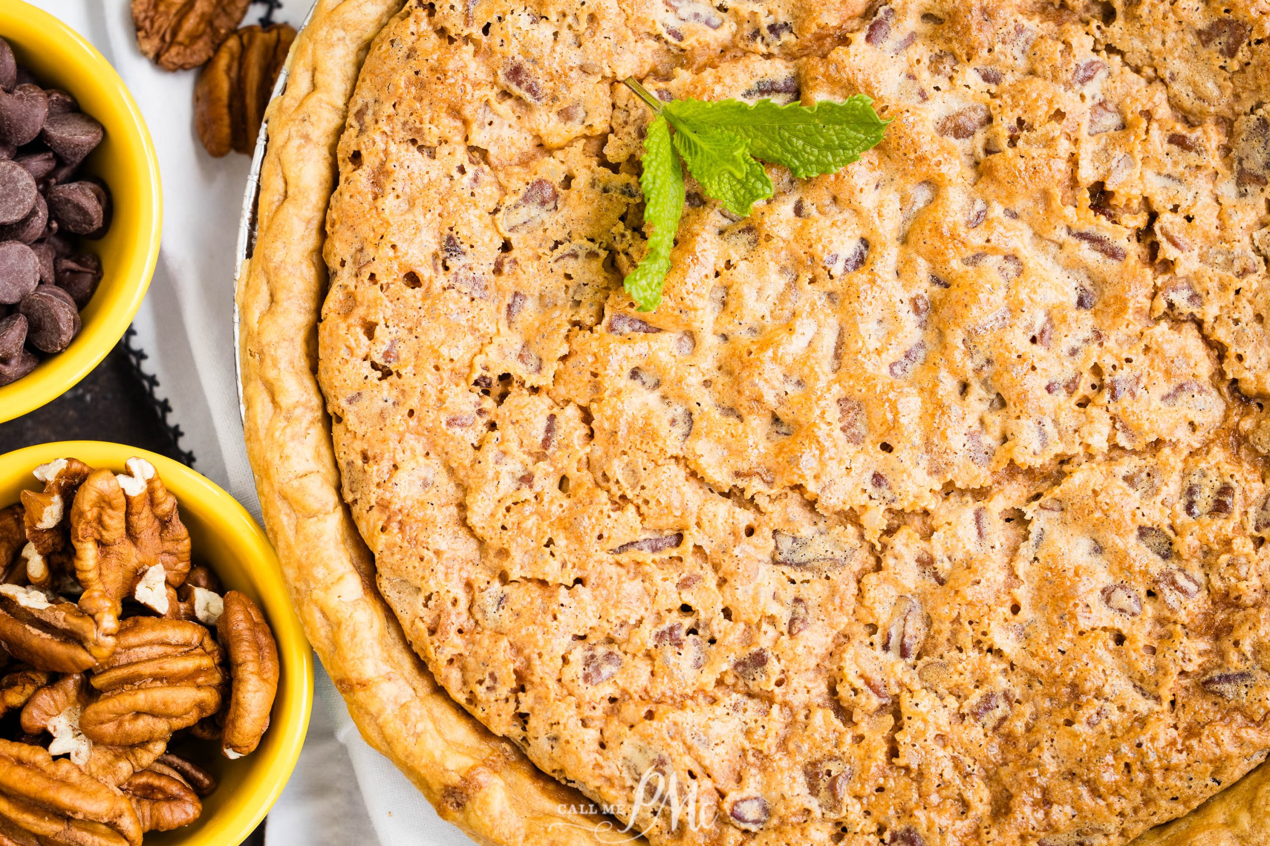 A whole pecan pie topped with a sprig of mint, accompanied by small bowls of pecans and chocolate chips.