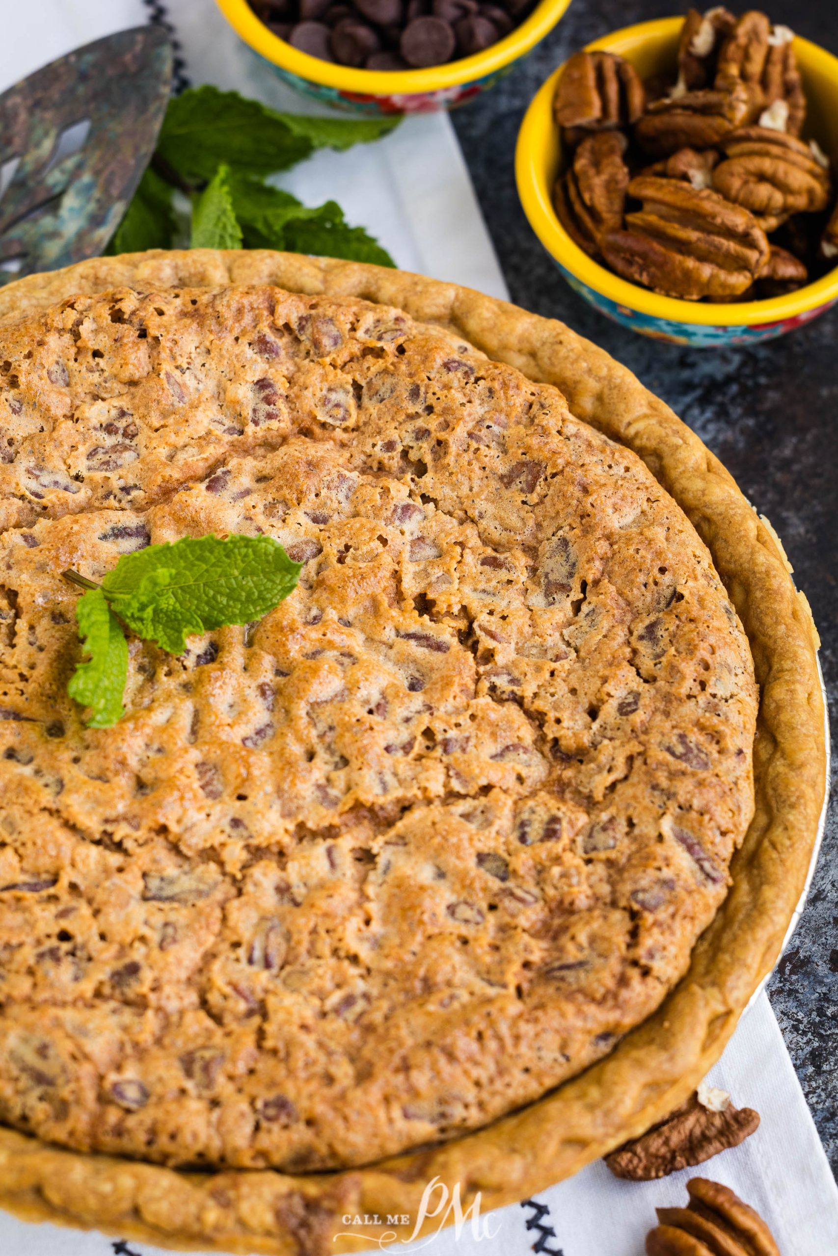 A Kentucky Derby Pie garnished with a mint leaf is placed on a table. Nearby, there are bowls containing chocolate chips and pecans.