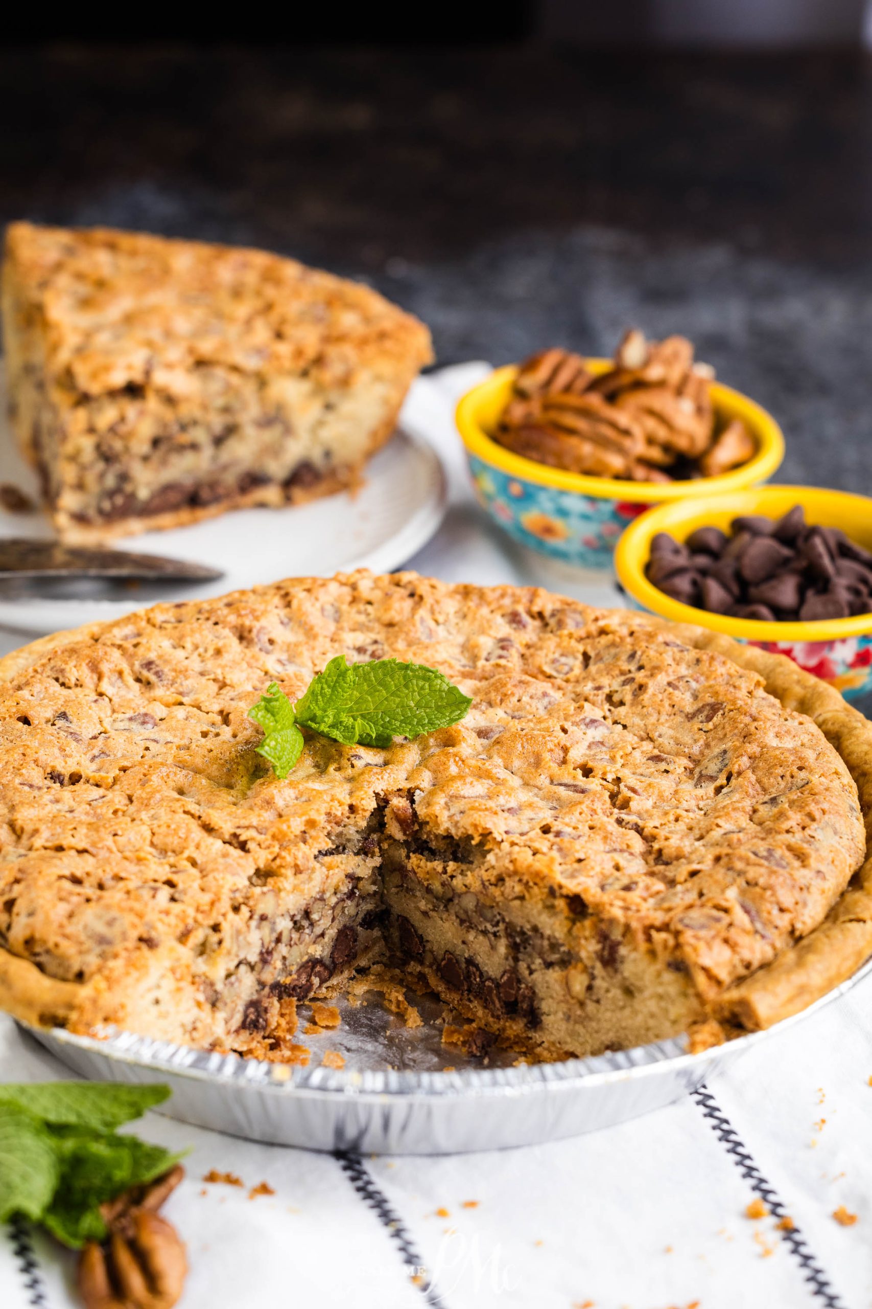 A pecan pie with a slice removed, revealing the inside with chocolate chips. The slice is placed on a white plate in the background. Small bowls of pecans and chocolate chips are nearby.