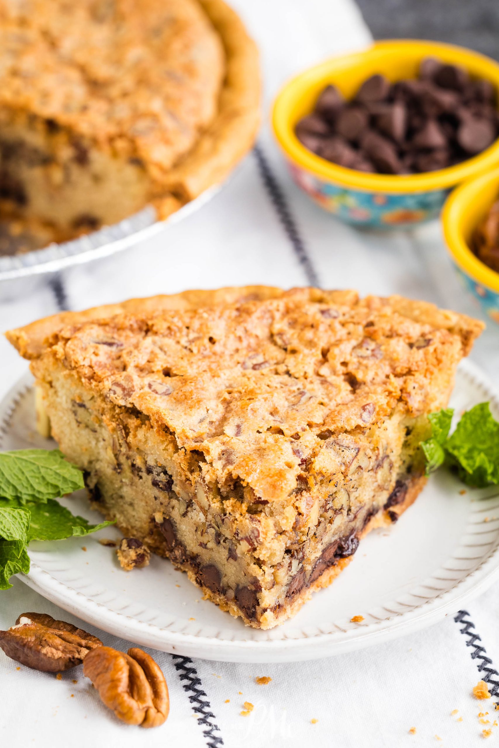 A slice of Kentucky Derby Pie with chocolate chips on a white plate, garnished with mint leaves. In the background, bowls of chocolate chips and whole pecans are visible.