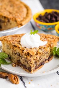 Slice of pecan pie with chocolate chips, topped with whipped cream and a mint sprig, served on a white plate.