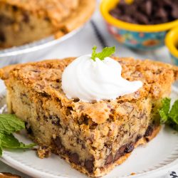 Slice of pecan pie with chocolate chips, topped with whipped cream and a mint sprig, served on a white plate.