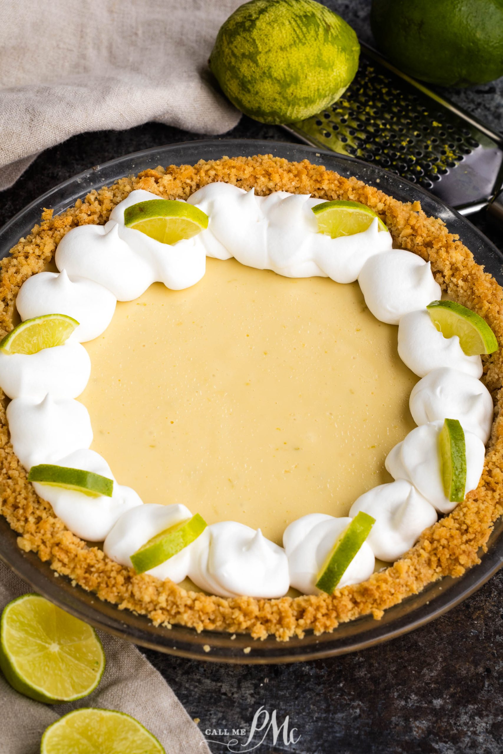 A key lime pie with a graham cracker crust, topped with swirls of whipped cream and lime slices, placed on a table with whole and cut limes in the background.