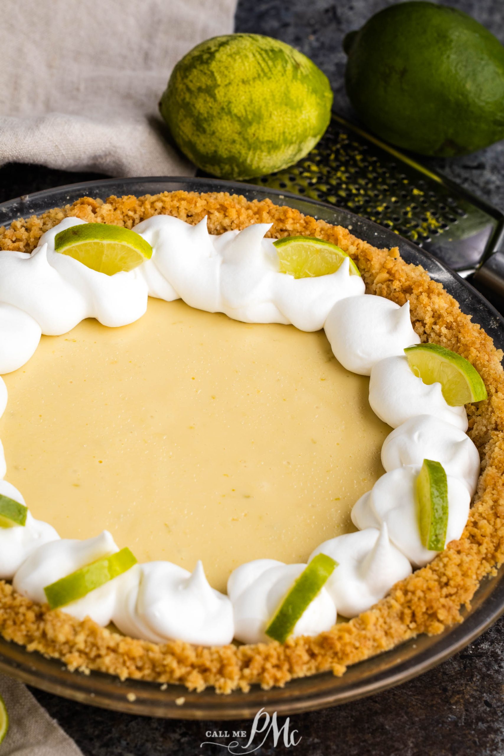 A dessert with a graham cracker crust, topped with whipped cream and lime slices, sits on a dark countertop. A zested lime and another whole lime are in the background.