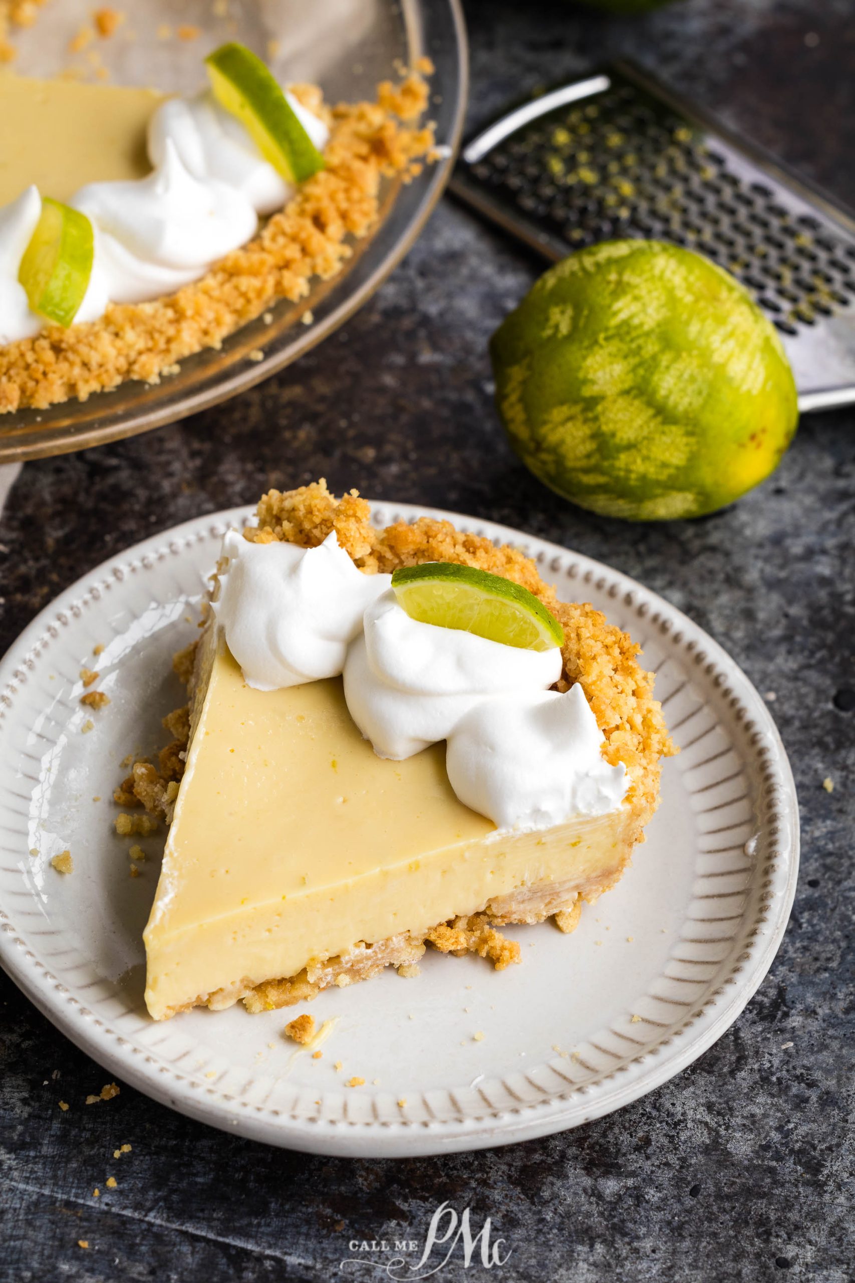 A slice of key lime pie on a white plate, garnished with whipped cream and a lime wedge. A pie dish with the remaining pie and a whole lime are in the background.