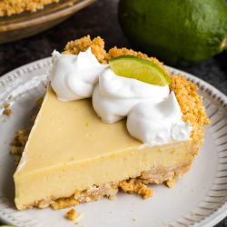 A slice of key lime pie sits on a plate, topped with whipped cream and a lime wedge. A whole pie and limes are visible in the background.