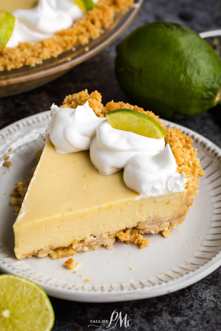 A slice of key lime pie sits on a plate, topped with whipped cream and a lime wedge. A whole pie and limes are visible in the background.