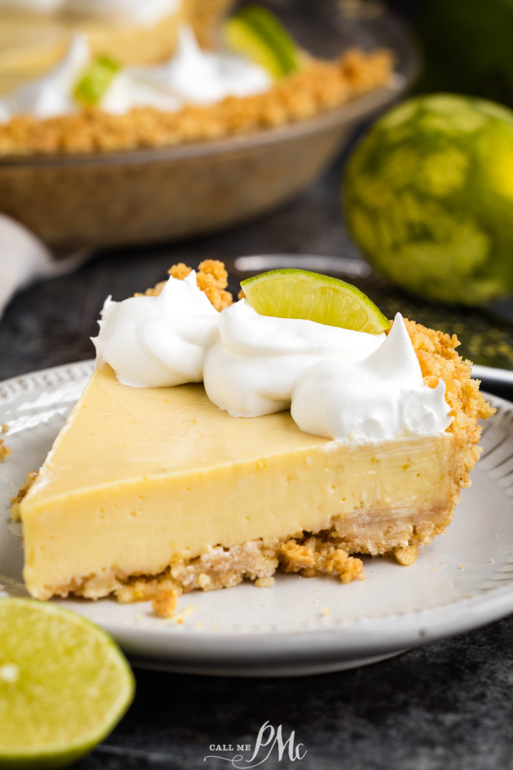 A slice of key lime pie topped with whipped cream and a lime wedge, sitting on a white plate with a blurred pie and lime in the background.