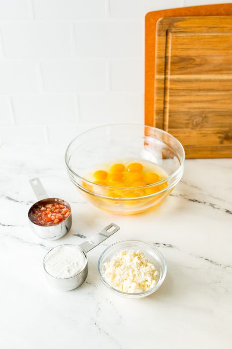 A glass bowl with cracked eggs, a measuring cup containing diced tomatoes, another with flour, and a small bowl with crumbled cheese are on a marble countertop near a wooden cutting board.