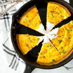 A cast iron skillet with a frittata cut into six slices, garnished with herbs and a dollop of sour cream, sits on a white and black plaid cloth next to two forks.