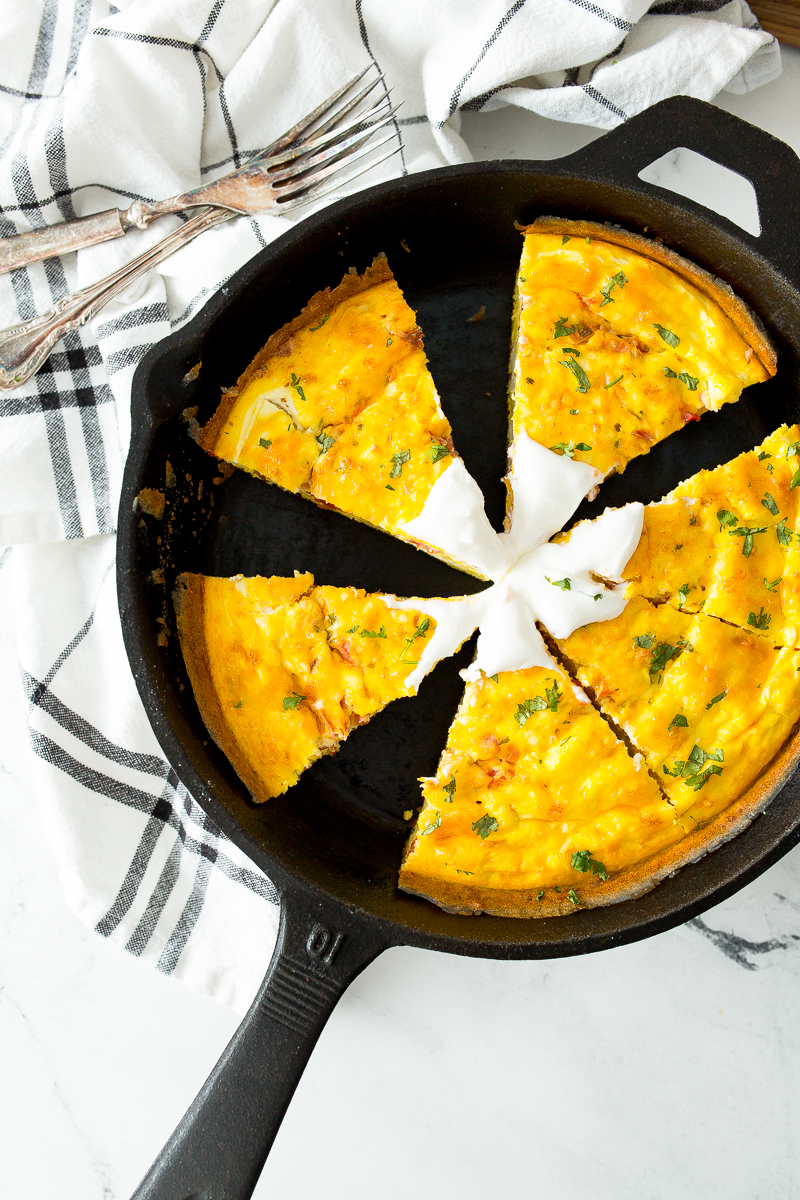 A cast iron skillet with a frittata cut into six slices, garnished with herbs and a dollop of sour cream, sits on a white and black plaid cloth next to two forks.