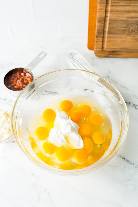 A glass bowl with several raw eggs and some dollops of white yogurt. A whisk, a measuring cup with salsa, and a wooden cutting board are nearby on a marble countertop.