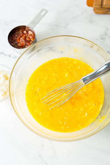 A glass bowl with a yellow liquid and whisk, a measuring cup with diced ingredients, and a small dish with white powder on a marble countertop.