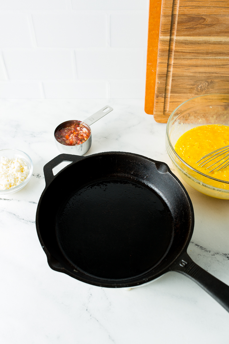 A cast iron skillet on a marble countertop, accompanied by a bowl of beaten eggs, diced vegetables in a measuring cup, grated cheese in a small bowl, and a wooden cutting board in the background.