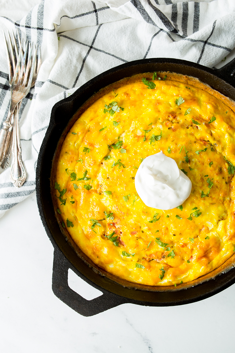 A cooked frittata in a cast iron skillet, garnished with herbs and a dollop of sour cream, rests on a white and black checkered cloth with three silver forks beside it.