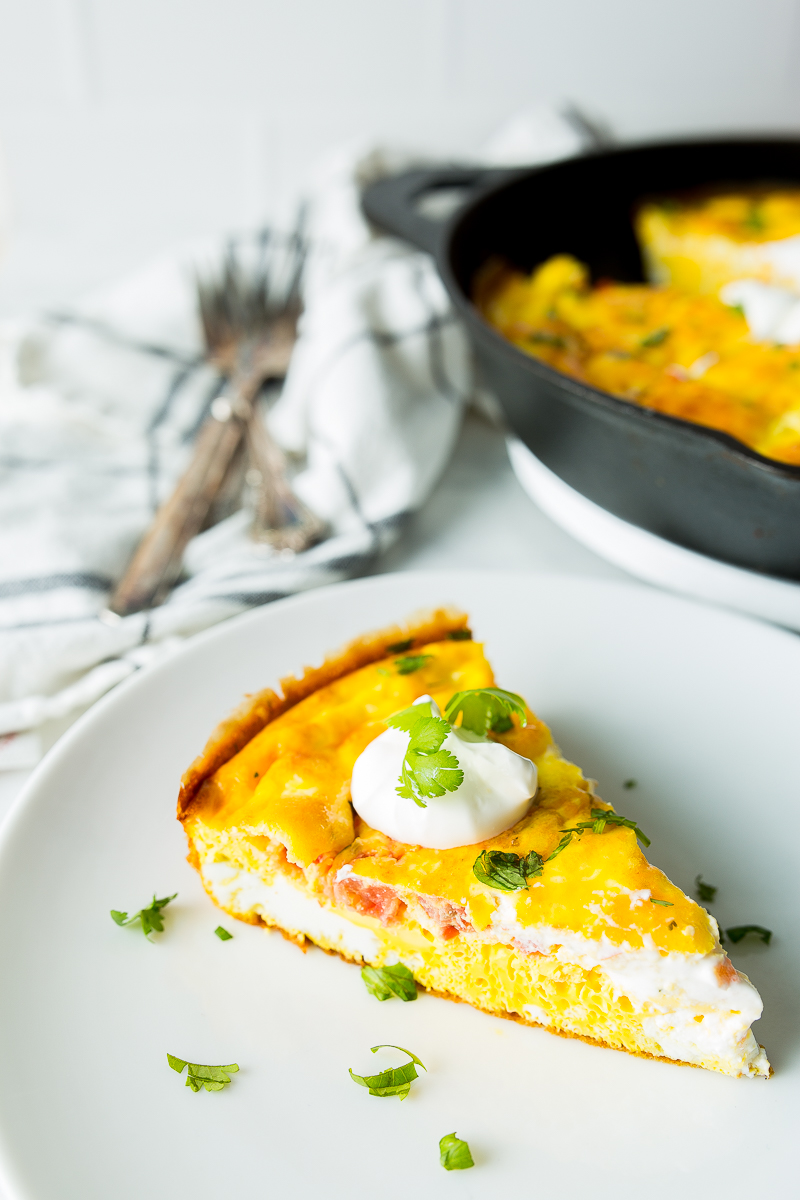 A slice of frittata topped with sour cream and garnished with cilantro on a white plate, with a pan containing more frittata and a napkin with forks in the background.