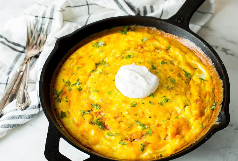 A Cast Iron Mexican Frittata sizzles in a black skillet, crowned with a dollop of sour cream and garnished with chopped herbs. A fork rests elegantly on a cloth napkin beside it.