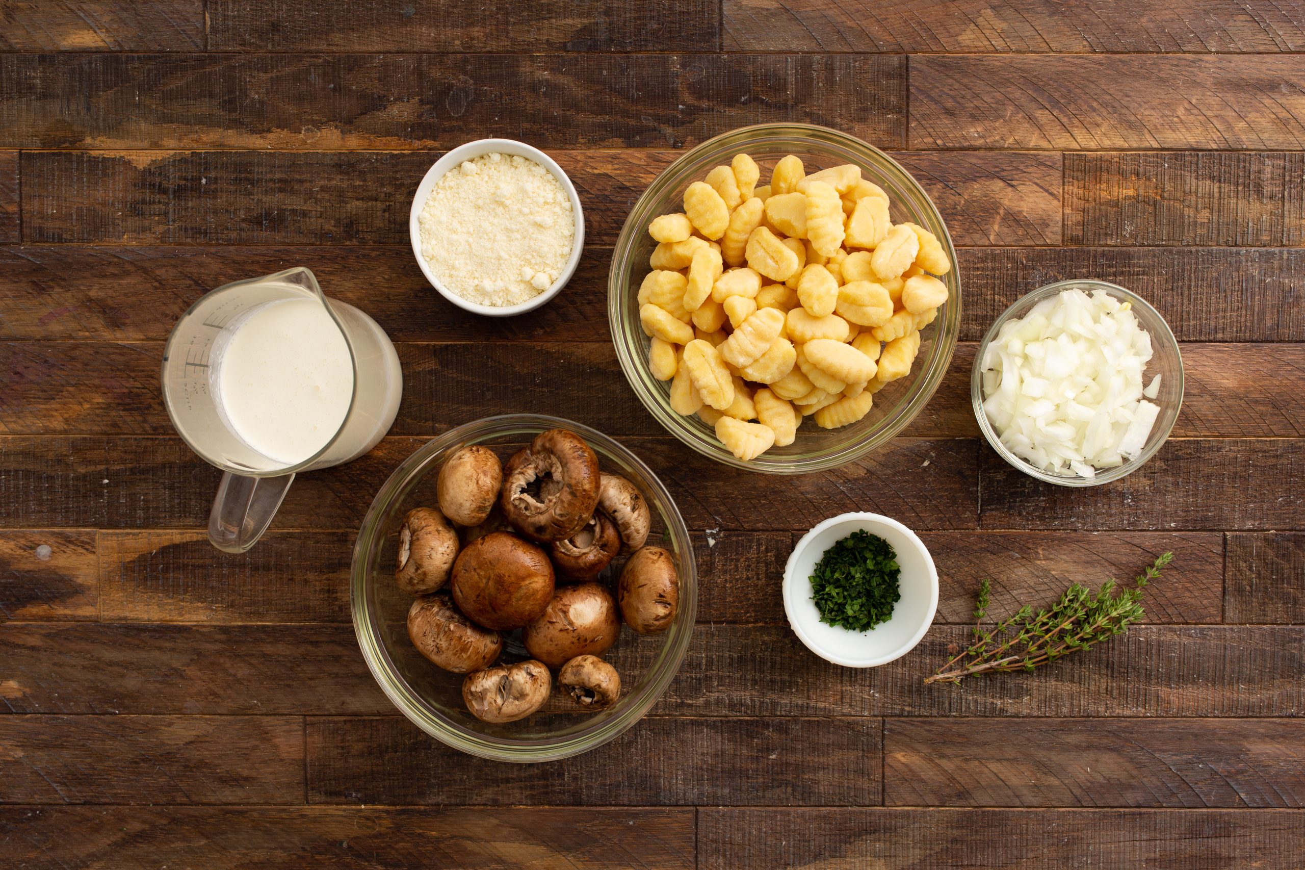 A wooden surface holds bowls of cream, grated cheese, gnocchi, diced onions, whole mushrooms, chopped parsley, and thyme sprigs arranged neatly.